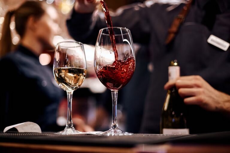 A bartender elegantly pouring red wine into a glass next to a glass of white wine at a bar, with a blurred background of a busy restaurant setting.