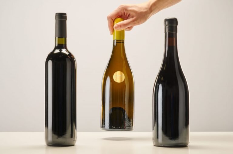 A hand lifting a premium white wine bottle with a yellow cap between two dark glass red wine bottles on a minimalist white background.