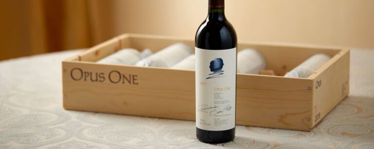 Luxury bottle of Opus One 2019 wine standing in front of a wooden case with other bottles inside.
