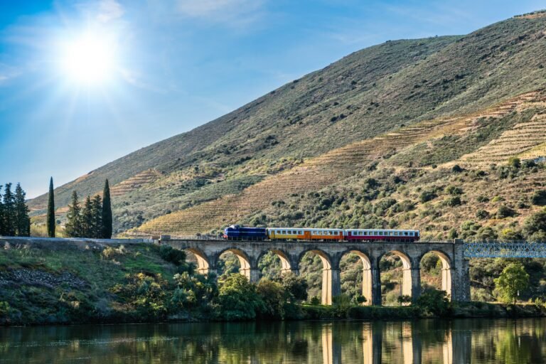 Colorful vintage train crossing a stone viaduct in the Douro Valley, Portugal, with terraced vineyards on green hills under a sunny blue sky.
