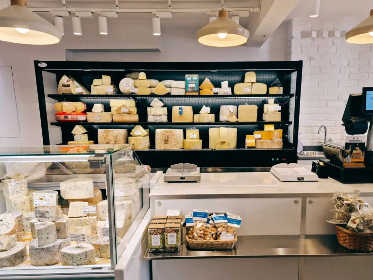A cheese counter filled with a wide range of artisan European cheeses, including blue, hard, and raw milk varieties, in a bright and modern cheesemonger’s shop.