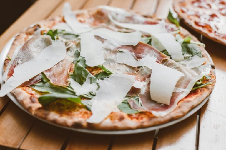 A wood-fired pizza topped with fresh rocket, slices of cured Parma ham, and generous shavings of Parmesan cheese, served on a wooden table.