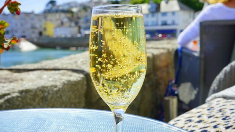 Close-up of a glass of sparkling wine on a sunny seaside terrace, with golden bubbles rising and a blurred coastal village in the background.