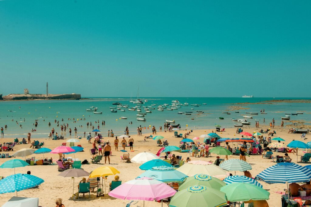 Crowded summer beach in Cádiz, Spain, filled with colourful umbrellas and sunbathers, with boats floating on turquoise water and a historic fort in the background.