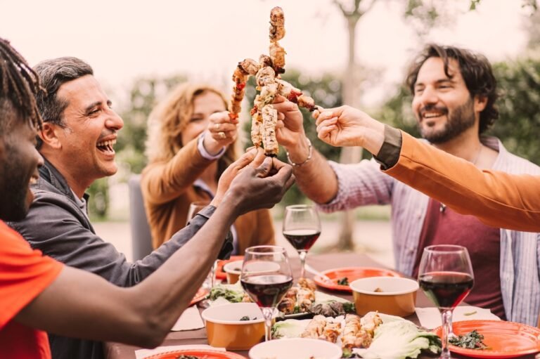 Friends enjoying barbecue skewers with glasses of red wine at a summer outdoor gathering.