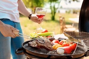Person grilling steak and vegetables on a barbecue while holding a glass of rosé wine outdoors.