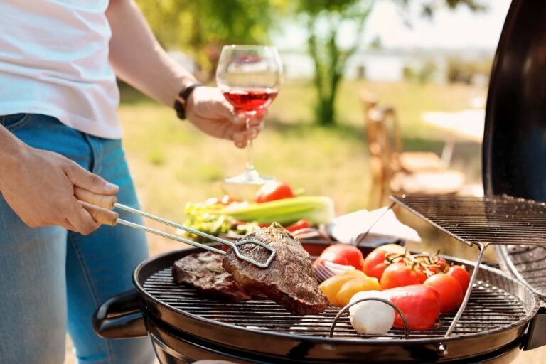 Person grilling steak and vegetables on a barbecue while holding a glass of rosé wine outdoors.