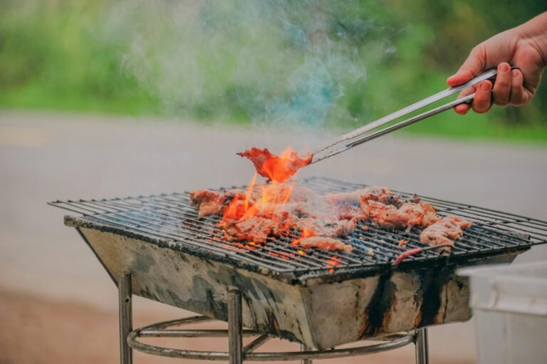 Outdoor barbecue with sizzling meat on a charcoal grill, flames and smoke rising as a hand flips meat with tongs