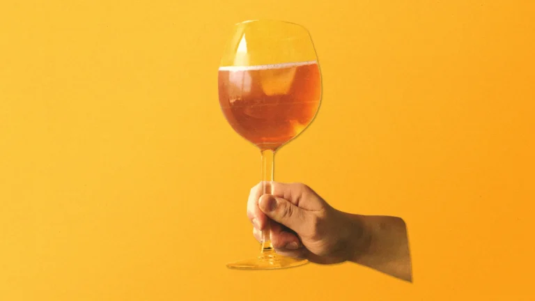 Hand holding a glass of orange wine against a vibrant orange background.