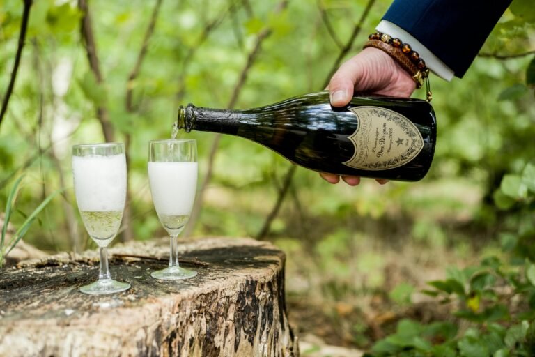 Hand pouring Dom Pérignon champagne into two glasses outdoors on a rustic wooden surface.