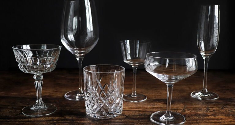 Different types of elegant glassware on a wooden table, including wine glasses, champagne flutes, and cocktail glasses under soft lighting.