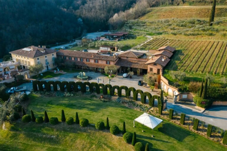 Aerial view of a beautiful vineyard estate with traditional Tuscan architecture, green vineyards, and manicured gardens on a sunny day.